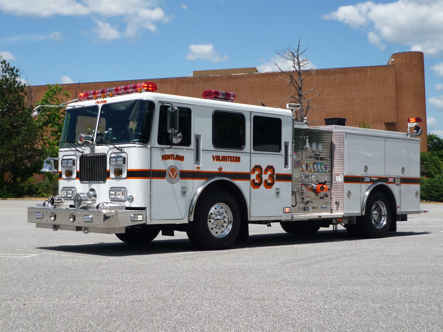 A white volunteer fire truck labeled "33" and "Kentland Volunteers" is parked on an empty paved lot with a brick building and green trees in the background under a partly cloudy sky.