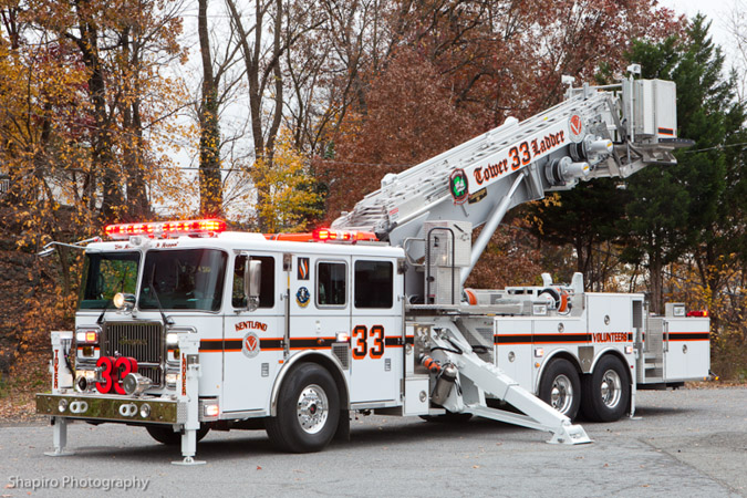 A white fire truck with red and black accents, labeled “33” and “Tower 33 & Ladder,” is parked outdoors. Its ladder is extended, and red emergency lights are on. Autumn trees with brown leaves are in the background.