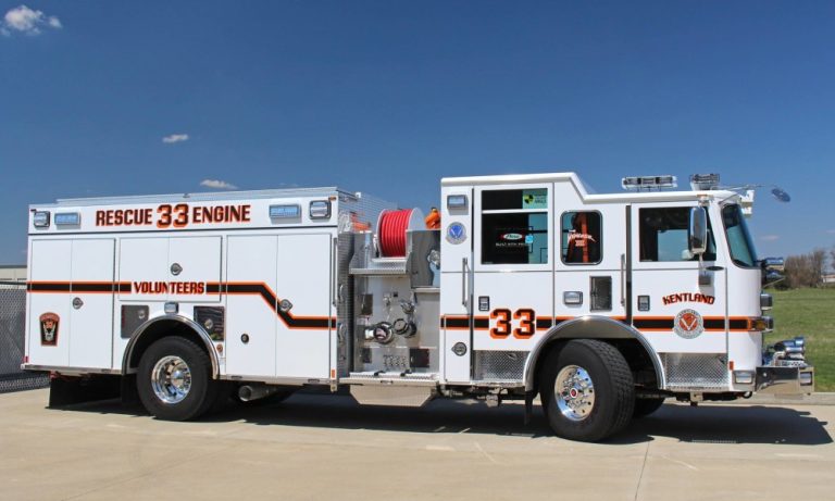 A white fire rescue truck labeled "Rescue 33 Engine" and "Volunteers" is parked on pavement under a clear blue sky. The truck has red and black stripes and "Kentland" written on the door.