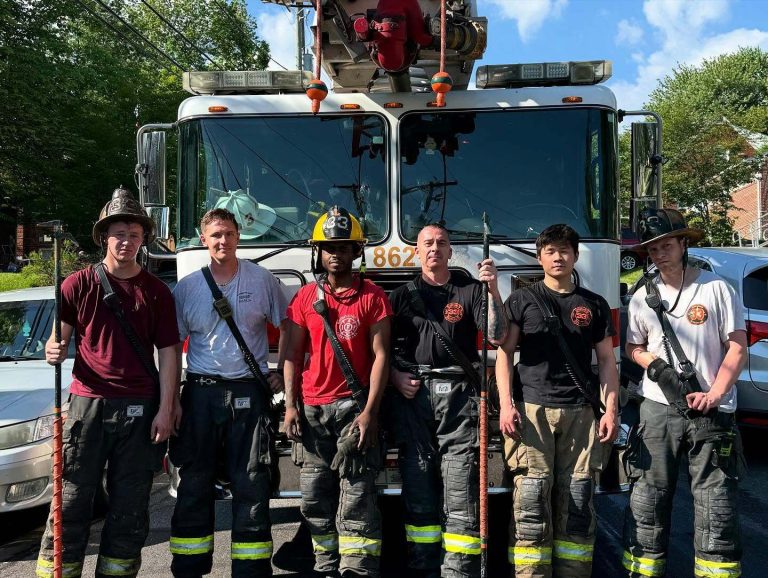 Six firefighters in uniform stand in front of a fire truck on a sunny day, holding firefighting tools and wearing helmets or hats, with trees and cars in the background.