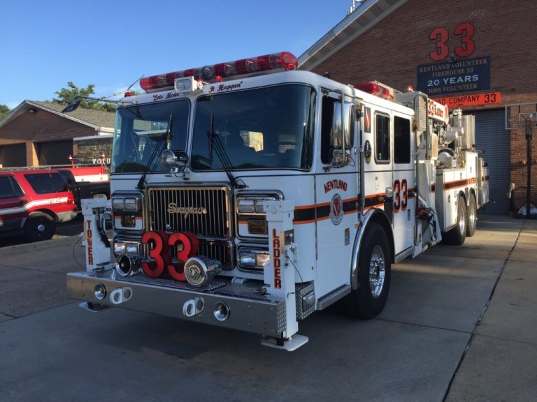 A large white fire truck with the number 33 on the front is parked outside a fire station. The building behind it also displays the number 33 and a sign about 20 years of service.