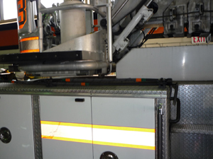 Close-up of a fire truck’s side compartments, showing metal storage panels and part of an extended ladder mechanism above. Reflective yellow and orange stripes are visible on the compartment doors.