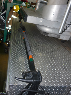 A firefighter in yellow gear stands on a metal diamond-plate surface next to a large piece of equipment, with a long black firefighting tool with orange markings laid out in the foreground.