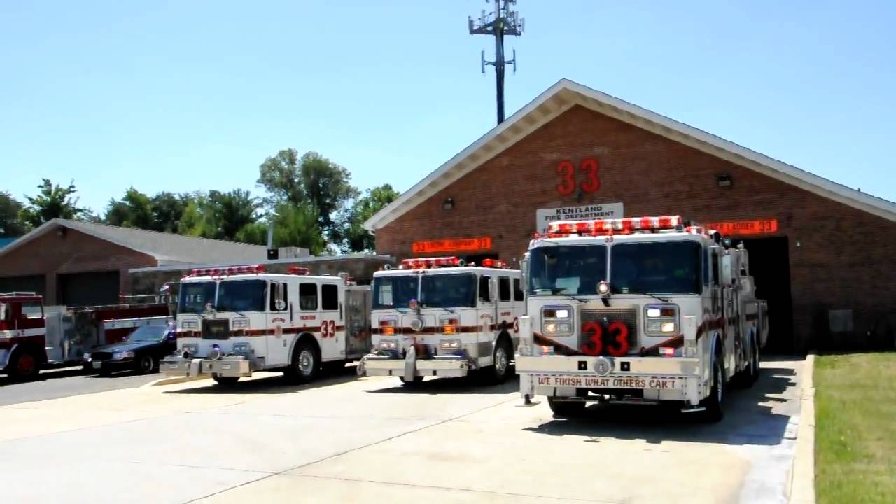Three fire trucks are parked outside a brick fire station with the number 33 displayed above the garage doors. The trucks have red and white markings, and the fire station is surrounded by trees and other buildings.