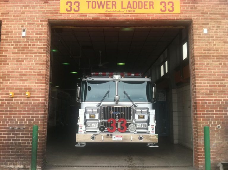 A fire truck labeled "33" is parked inside a brick fire station bay, beneath a sign that reads "Tower Ladder 33" with "Established 1988" above the entrance.
