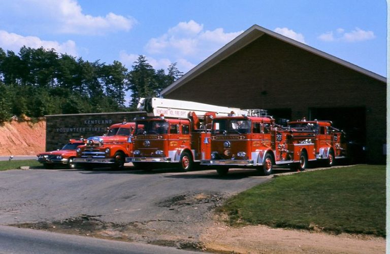 Four vintage red fire trucks are parked outside a brick fire station with an open garage door. A red car is also visible. The sign on the building reads "Kentland Volunteer Fire Dept." Trees and blue sky are in the background.