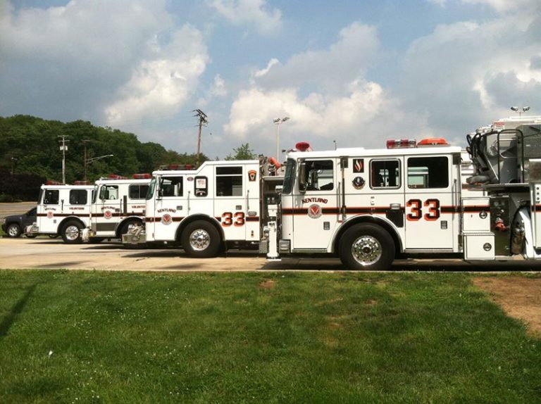 Four white fire trucks with red stripes and the number 33 are parked in a row on a paved lot beside a grassy area, with trees and a cloudy sky in the background.