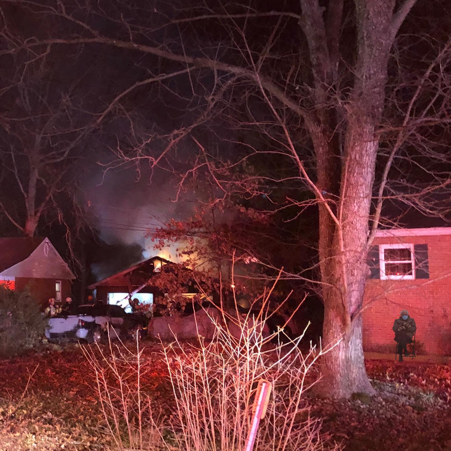 A house is on fire at night, with flames and smoke visible. Bare tree branches and two nearby houses are seen in the foreground. Emergency responders are present to the right.