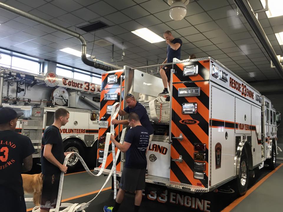 Four firefighters are inside a fire station, unrolling and organizing hoses at the back of a fire engine labeled "Rescue 33 Engine." Another fire truck is visible in the background.