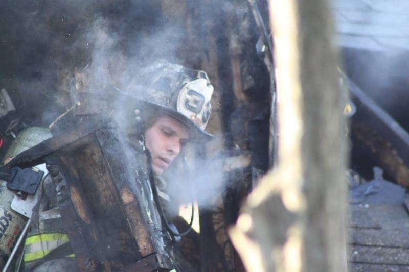 A firefighter wearing a helmet is partially obscured by smoke while working amidst charred debris, showing determination and focus during an emergency response.