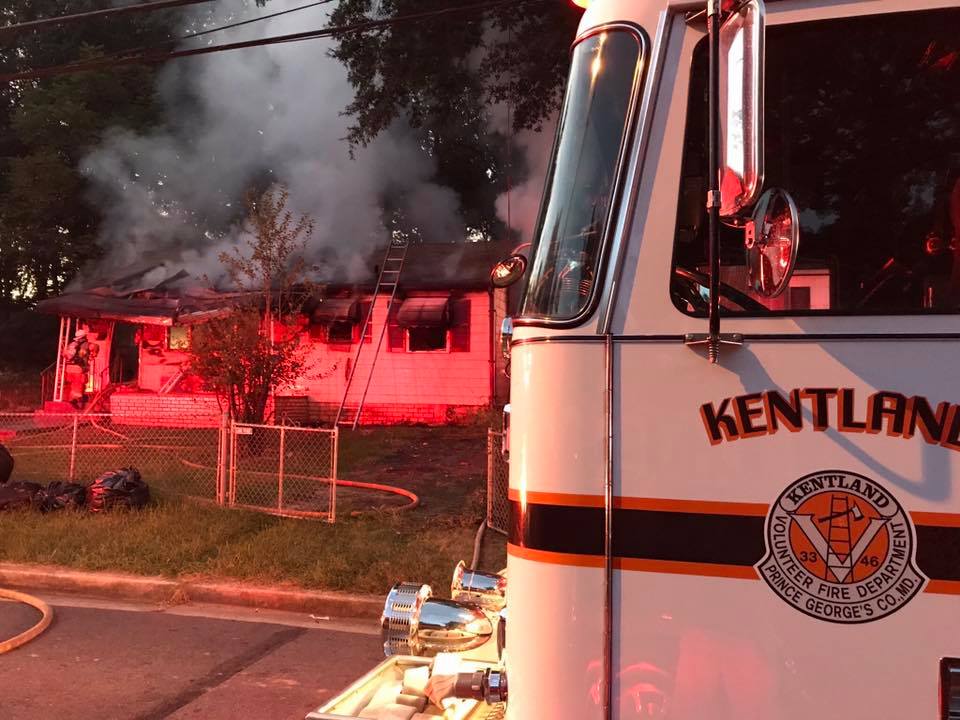 A fire truck labeled "KENTLAND" is parked in front of a single-story house with smoke rising from its roof as firefighters respond to a fire. Red emergency lights illuminate the scene at dusk.