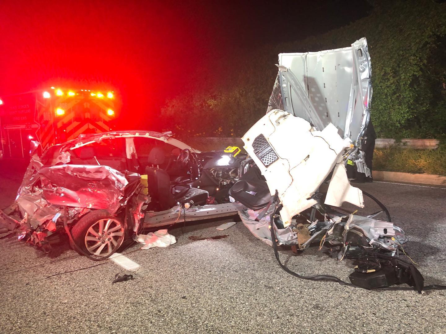 A heavily damaged silver car with its sides and front severely crushed sits on a road at night. Emergency vehicle lights are visible in the background, illuminating the scene.