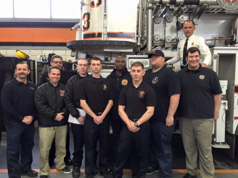 A group of men, some in black uniforms and others in casual clothing, pose for a photo inside a fire station. One man, wearing white, stands elevated beside fire equipment in the background.