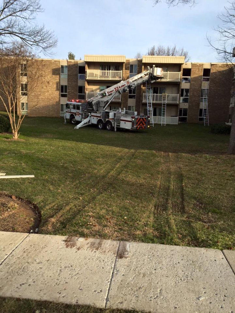 A fire truck with an extended ladder is parked on the grass in front of a three-story apartment building, leaving deep tire tracks on the lawn. The weather appears clear and the trees are leafless.