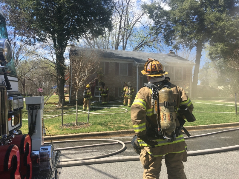 A firefighter in full gear stands near a fire truck, watching as other firefighters respond to a house fire. Smoke rises from the house as hoses are laid out on the lawn and driveway. Trees surround the scene.