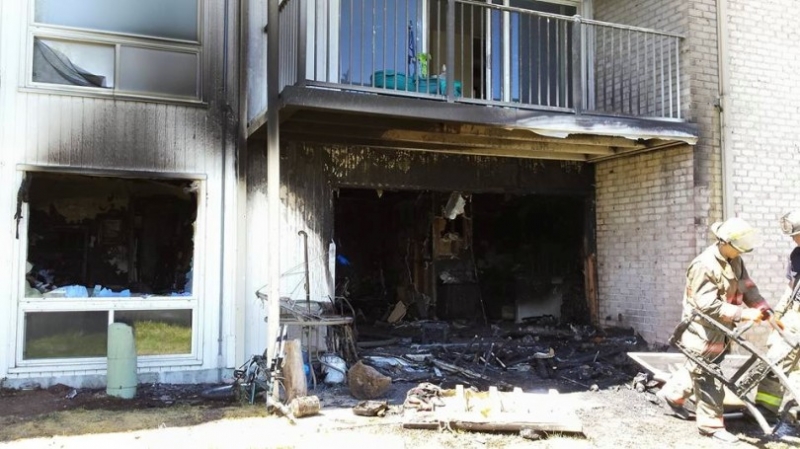 A burned apartment complex with blackened walls, broken windows, and debris scattered outside. Two firefighters in gear are at the scene, assessing damage and working near the charred remains.