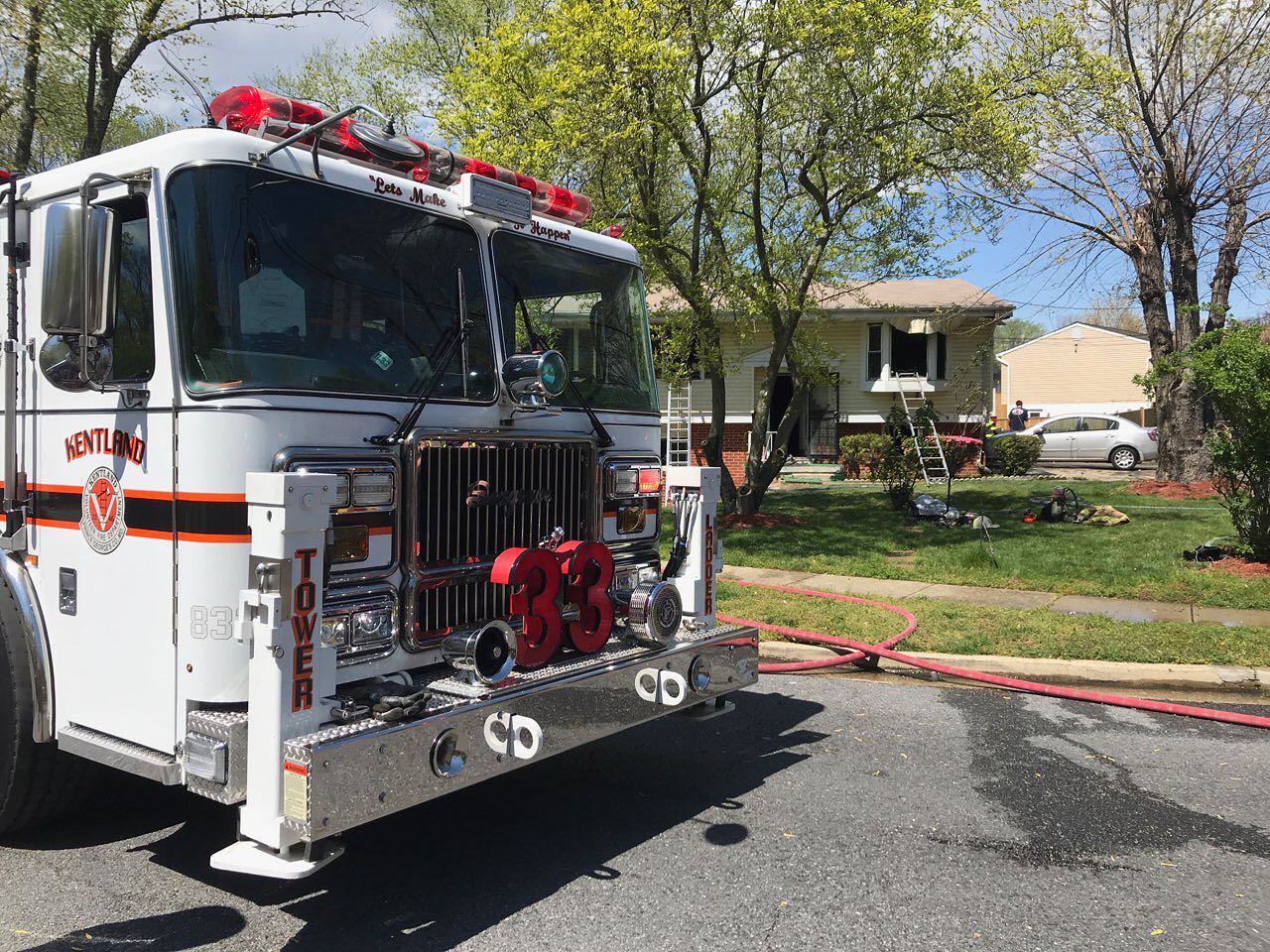 A fire truck labeled "33" is parked in front of a house with an open window and fire damage. Ladders are set up to the window, and fire hoses are spread across the lawn. Trees and blue sky are in the background.
