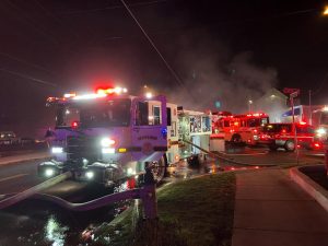 A dark car with severe rear-end damage sits on a road at night. Emergency vehicles, including a Rescue 33 fire engine, are present with flashing lights, and responders are at the scene.