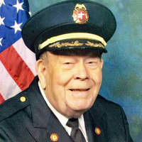 Elderly man in a formal firefighter's uniform and cap, smiling, with an American flag in the background.