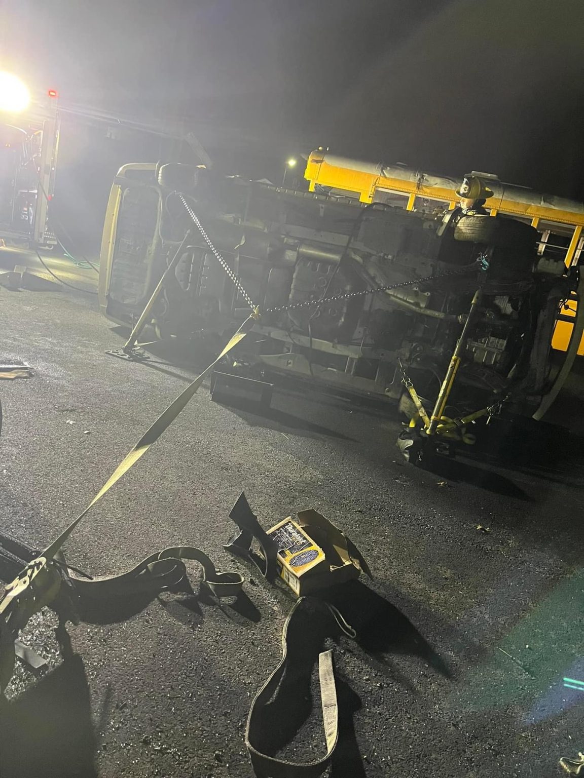 A yellow school bus lies on its side at night on a paved surface, with recovery straps attached. Emergency lights illuminate the scene, and equipment is scattered on the ground.
