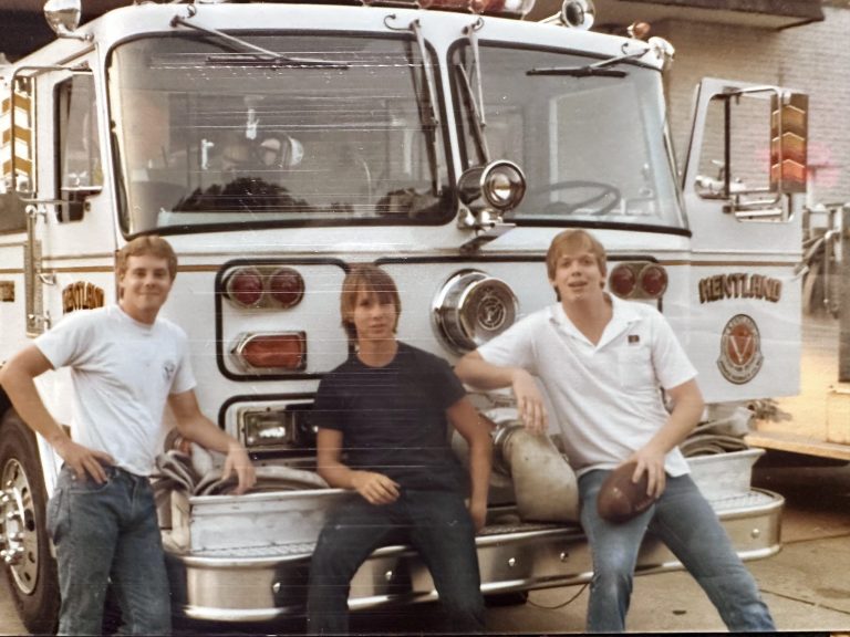 Three young men pose in front of a white fire truck. Two stand on either side, and one sits on the bumper. The man on the right holds a football. All three are dressed casually in jeans and t-shirts.