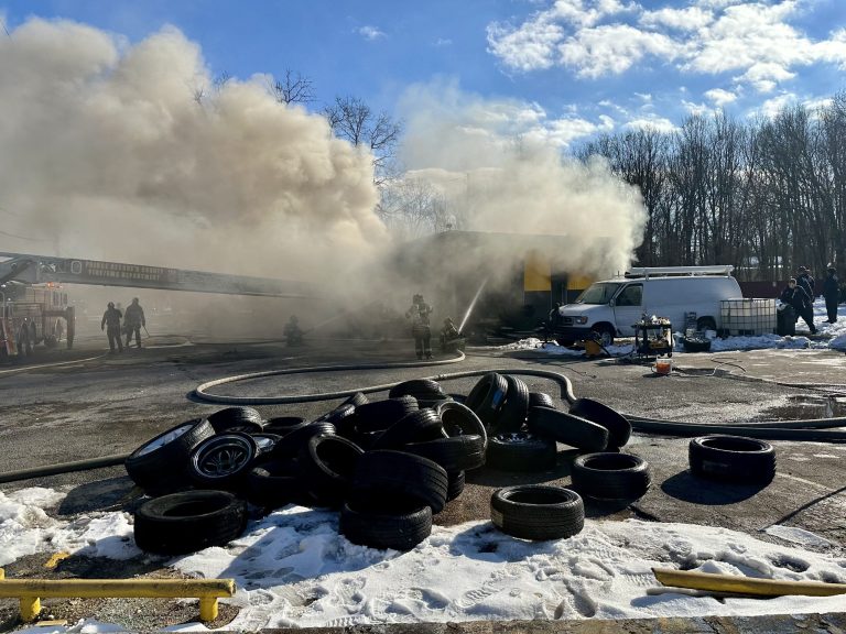 Firefighters work to extinguish a smoky fire at a building, with hoses spraying water. A pile of tires, snow patches, and a white van are in the foreground, while smoke billows into the sky under scattered clouds.
