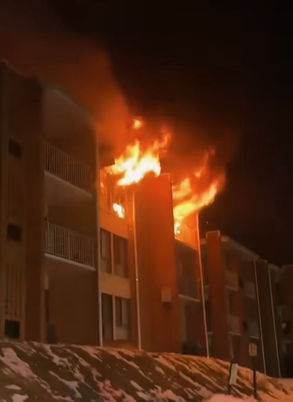 A large fire engulfs the upper floor and roof of a multi-story apartment building at night, with flames and smoke pouring out. Snow covers the ground in front of the building.
