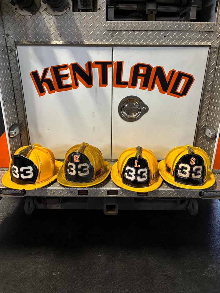 Four yellow firefighter helmets labeled "33" are lined up on the back bumper of a fire truck with "KENTLAND" written in orange and black letters above them.