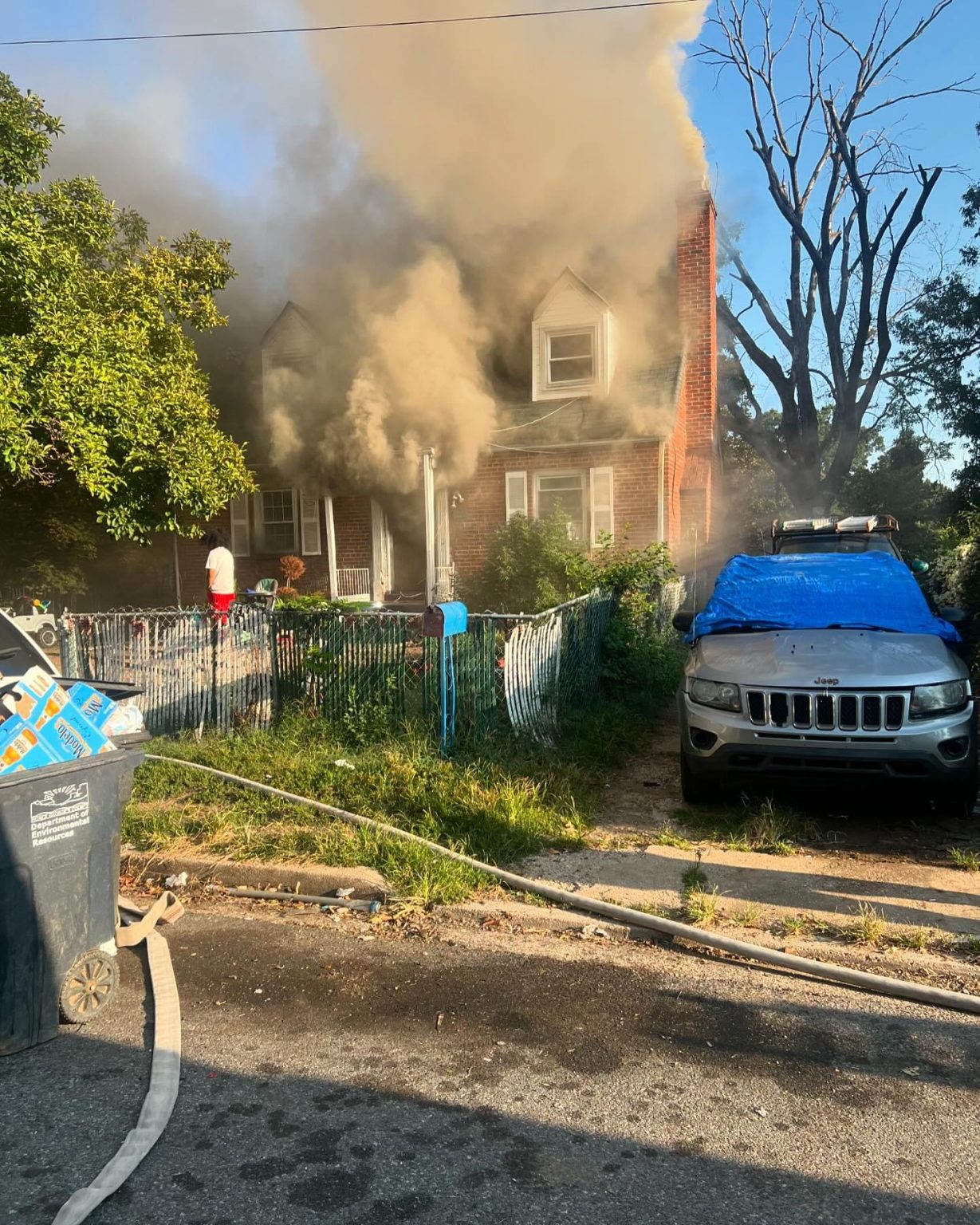 Smoke billows from the front of a house while a person stands on the porch. A nearby car is parked in the driveway, partially covered by a blue tarp, and a garden hose lies on the street.