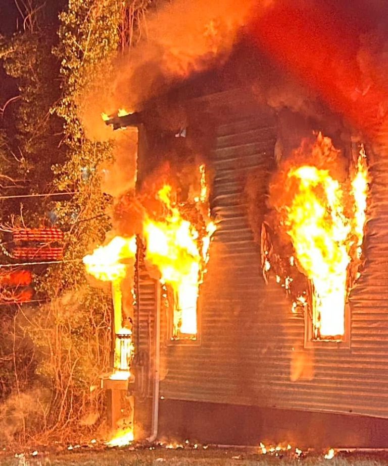 A house is engulfed in flames at night, with fire pouring out of multiple windows and thick smoke rising into the air. Trees and power lines are visible nearby.