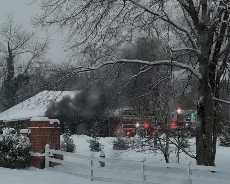 A fire truck with flashing lights is parked in front of a house as thick smoke billows from the building; snow covers the ground, trees, and fence, creating a wintery scene.