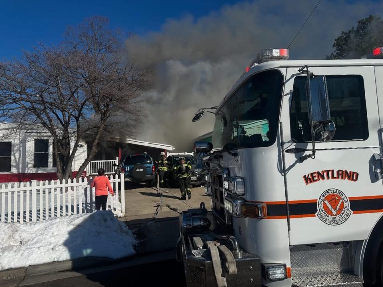 A fire truck labeled "Kentland" is parked near a white house with smoke billowing out. Firefighters in gear approach the house, while a person in a pink sweater stands outside a white picket fence. Snow covers parts of the ground.