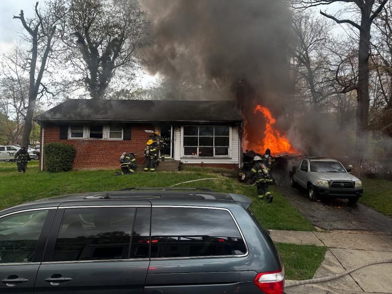 A house is on fire with large flames and smoke coming from a window. Firefighters in gear are responding on the front lawn and driveway, with cars parked nearby.