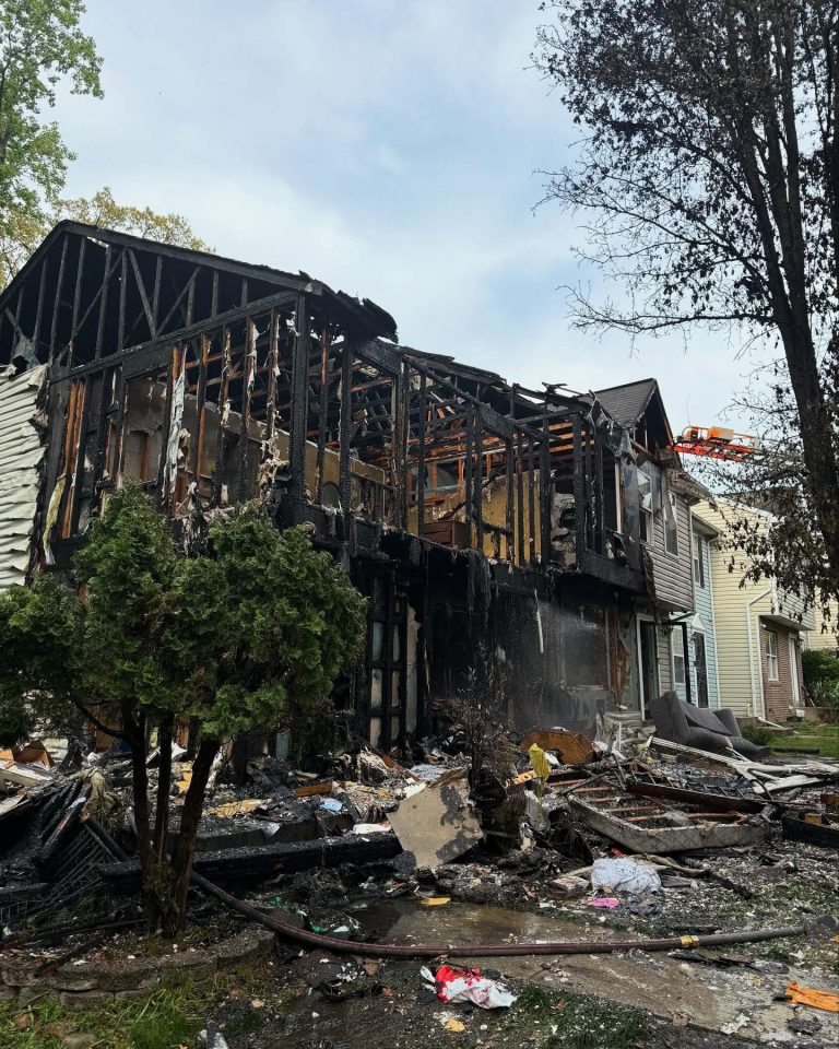 A severely burned two-story house with charred walls and debris scattered in the yard, showing extensive fire damage. Nearby trees and neighboring houses appear intact.