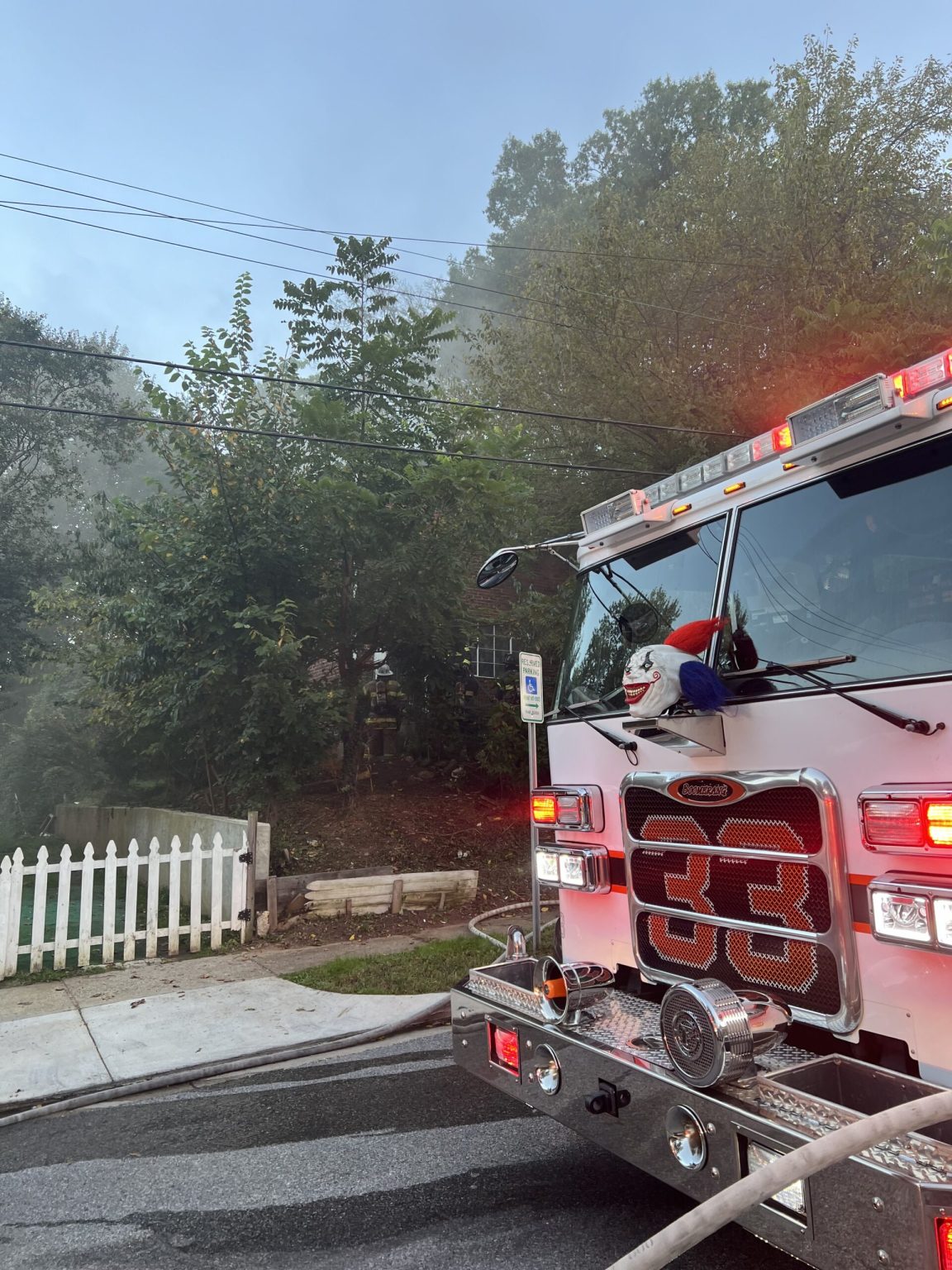 A fire truck with flashing lights is parked on a residential street near a white picket fence, with smoke rising from trees and a house in the background. A hose extends from the truck toward the scene.