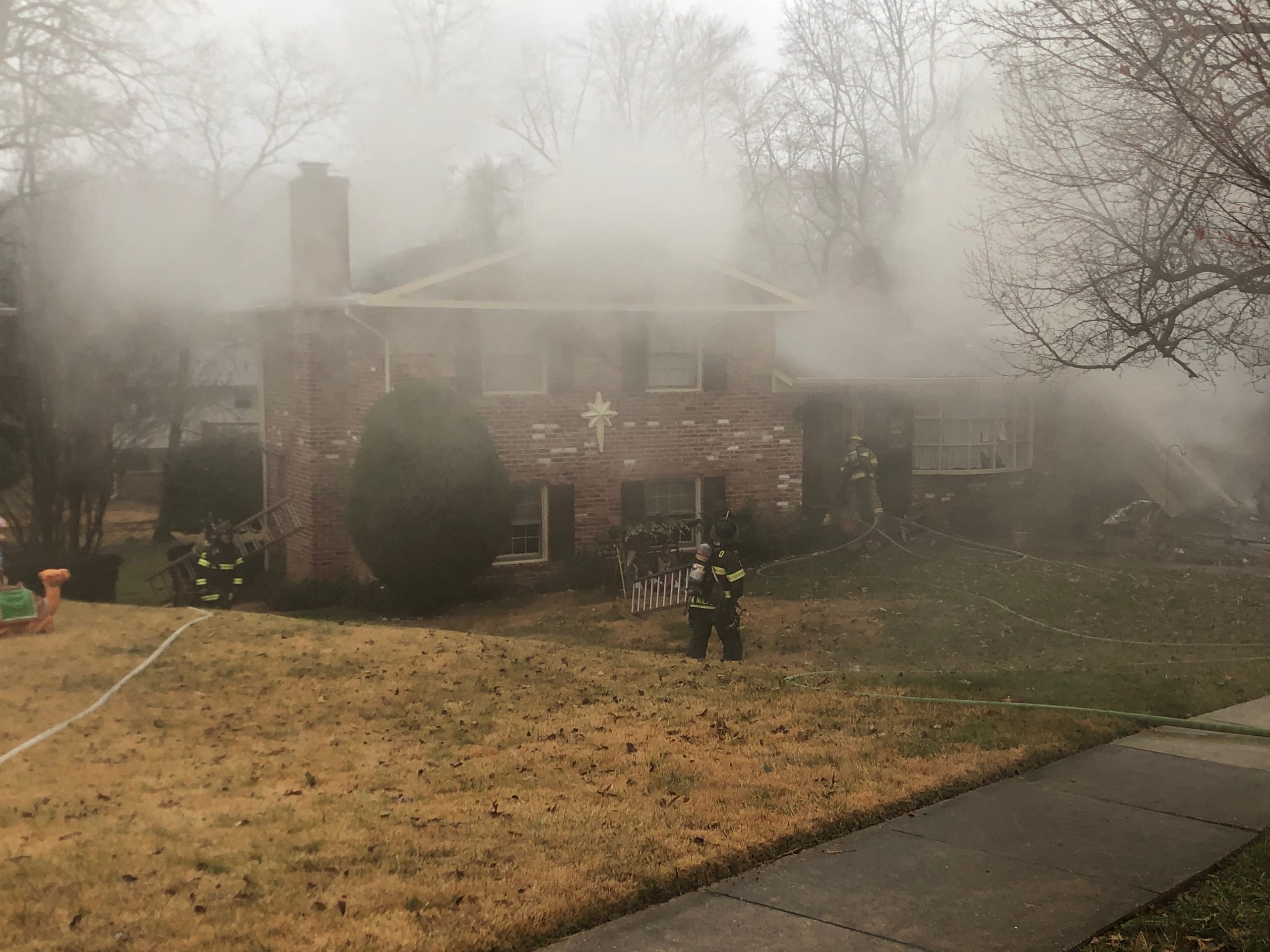 Four firefighters in gear stand in front of a fire truck on a residential street, with trees and houses in the background. Three wear helmets labeled “33,” and the fire truck’s ladder is visible above them.