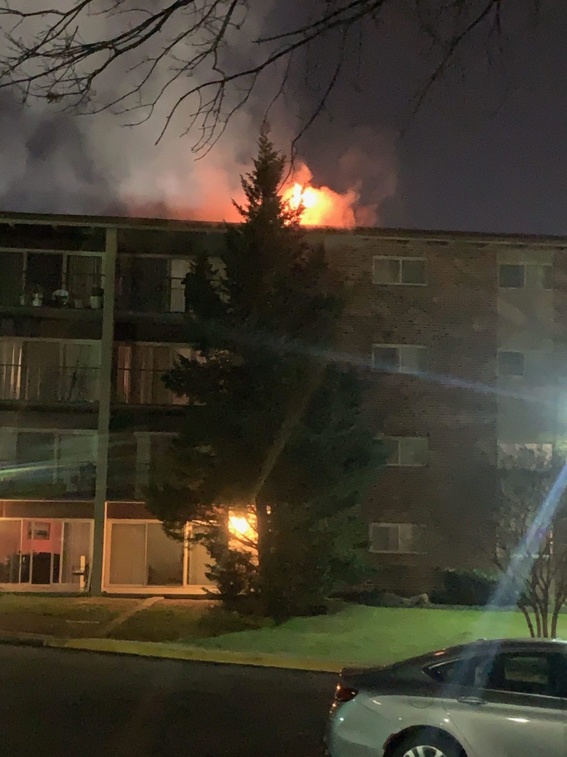 Flames and smoke are rising from the roof and a lower window of a brick apartment building at night, partially obscured by a tall tree. A parked car is visible in the foreground.