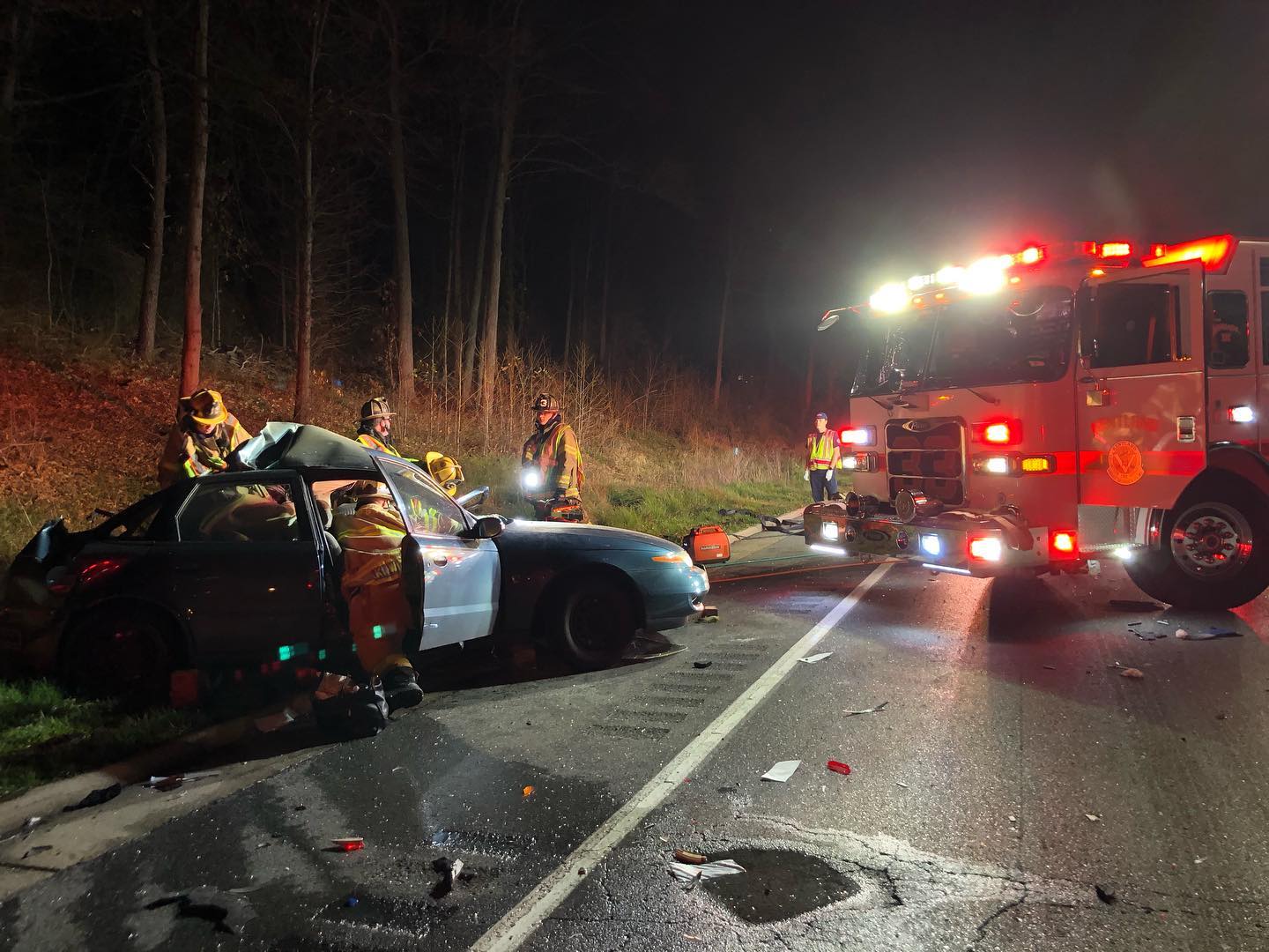 Firefighters respond at night to a car crash; a heavily damaged car is on the roadside with emergency lights flashing, and rescue workers attend to the scene near a fire truck.