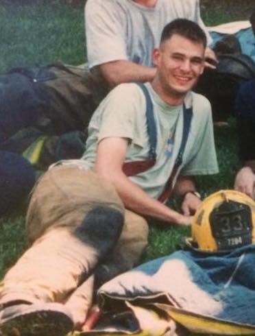 A young firefighter wearing suspenders, a t-shirt, and dirty turnout pants lies on the grass smiling, holding a yellow helmet with the number 33 on it. Other firefighters are sitting nearby.