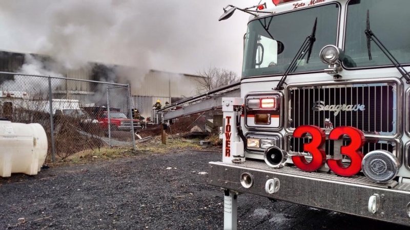 A fire truck labeled "33" is parked near a smoky industrial building. Firefighters are using a ladder and hose to combat the fire while smoke billows from the structure in the background.