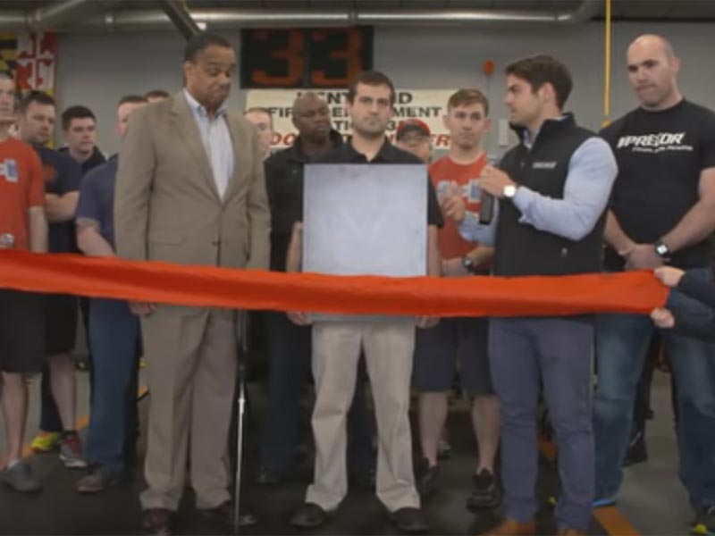 A group of people stand behind a red ribbon at an indoor event, with one man holding scissors as if preparing to cut the ribbon, while others watch and gather around.