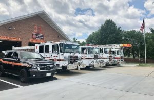 Four fire trucks and a black SUV are parked outside a brick fire station labeled "33" with an American flag on a pole and trees in the background under a partly cloudy sky.