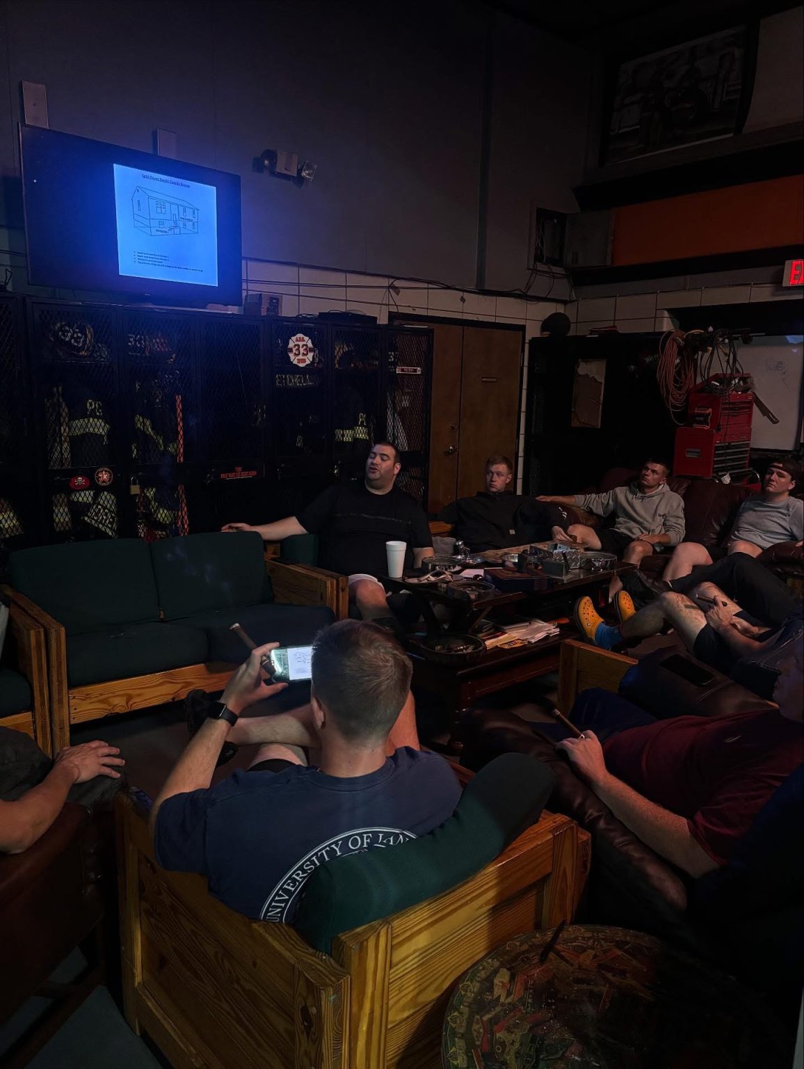 A group of people sit on wooden couches in a dimly lit room, watching a presentation on a wall-mounted TV. Firefighting gear is visible in the background, and one person uses a smartphone.