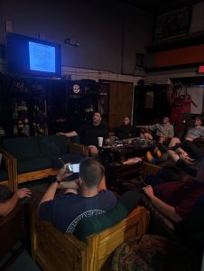 A group of people sit on wooden couches in a dimly lit room, watching a presentation on a wall-mounted TV. Firefighting gear is visible in the background, and one person uses a smartphone.