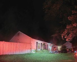 A nighttime scene shows firefighters entering a single-story house with smoke visible near the roof. Red emergency lights illuminate the house, and trees and a wooden fence are nearby.