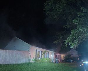 Nighttime scene of a single-story house with smoke rising from the roof; a firefighter in gear stands near the front door. A large tree and a wooden fence are visible, with emergency vehicle lights illuminating the scene.