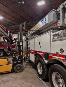 A forklift lifts part of a large white fire truck with red stripes inside a garage. Another fire truck is visible in the background under the building's high, exposed ceiling.