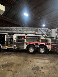 A fire truck with its side compartments open is parked inside a large building with a high, exposed ceiling. The truck's ladder is visible on top, and various equipment is stored inside the compartments.