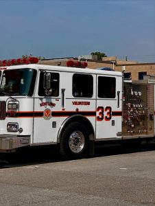 A white fire truck labeled "Kentland Volunteers 33" is parked on a street beside a brick building under a clear blue sky.