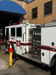 A white fire truck labeled "33 Volunteers" is parked outside a brick building with its side compartment open, displaying hoses and gauges. A stop sign and orange traffic cone are visible next to the truck.